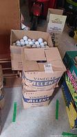 Four cardboard boxes stacked, containing golf balls. Boxes are worn with some damage and discoloration.