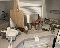 Photo of kitchen countertop with manual meat grinder, strainers, Bakerstone wooden cutting board, metal cups, rolling pin, pepper grinder, meat slicer, metal tray, ice cube mold, and plastic food grinder.