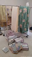Wide angle photo of rack with hanging and folded linens including sheets, drapes, and fabric pieces. Boxes of sheet sets and pillowcases on floor in front.