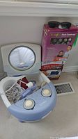 Ott-lite mirror with circular illuminated area, Homedics foot spa with control knobs and attachments inside basin, Dr. Ho's Neck Comforter and Pain Therapy System boxes, and FitOver sunglasses on top of boxes.