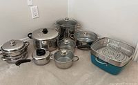 Overall view of all stainless steel pots, pans, glass and metal lids, including a blue enamel steamer pot on the right, showing all items included in the lot.