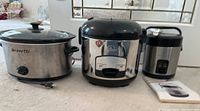 Three small kitchen appliances displayed: Bravetti 4 qt oval slow cooker, Wolfgang Puck model BDRC0007C rice cooker, and Wolfgang Puck 1.5 cup portable rice cooker with manual.