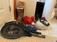 Photo showing two blue and black Spalding tennis rackets with one red boxing mitt, white helmet with goggles, jump rope, tennis balls, rubber baseball, and metal bat in background