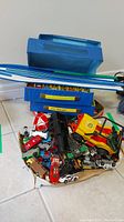 Loose pile of assorted toy cars, blue track rails, and blue carrying cases.