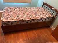 Single wooden bed frame with floral-patterned mattress seen from one angle, showing headboard and storage drawers under bed.