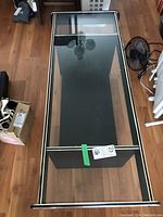 Photo of glass top coffee table with visible black wooden base and silver metal trim on edges, placed on wooden floor.