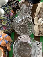Photo shows two clear crystal glass baskets with handles, four colorful floral and bird patterned ceramic pitchers behind them, a clear glass relish bowl next to the baskets, and two carved bird sculptures to the right.
