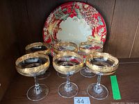 Six gold rimmed champagne glasses arranged with one decorative platter behind them on a wooden shelf.