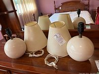 Pair of matching round cream-colored table lamps with accompanying beige conical lampshades with brown trim, placed on dresser in bedroom with mirror reflecting back.