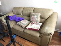 Front angle of beige leather couch with three decorative pillows and purple plush toy on the seat, showing wear on cushions and armrests.