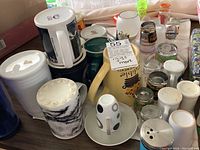 Photo showing multiple mugs, shot glasses, and small glassware on a table including a large beige mug with bee design and a marbled mug.