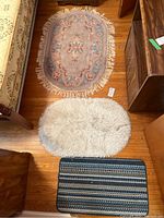 Three rugs on hardwood floor: oval pastel floral rug with tassels, oval cream shaggy mat, and small rectangular striped mat