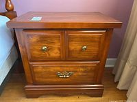 Front view of one wooden bedside table featuring two small upper drawers with round metal knobs and one large bottom drawer with an ornate handle, standing on a carved base.