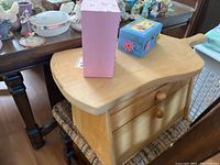 Wooden breadbox with two drawers shown from top angle, placed on a woven chair seat with pink and blue boxes on top.