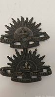 Front and back views of two WW2 Australian military pins. The pins feature the Australian Commonwealth Military Forces sunburst emblem with a crown and text. One has a rectangular clip back, the other has a safety pin back.