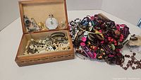 Wooden box filled with assorted costume jewelry and a pile of colorful beaded necklaces and bracelets beside it.