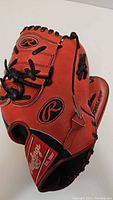 Angle showing the front and wrist strap area of the red Rawlings baseball glove with black laces and logo patches.