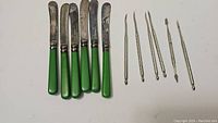 Photo shows 6 luncheon knives with green handles alongside 6 metal nut picks with decorative handles.