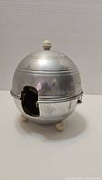 Side view of the vintage metal teapot warmer showing the front opening and three round feet for stability.