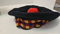 Top angled view of black wool tam hat showing red pom pom and striped knit band