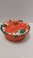 Top view of ceramic bowl with lid, decorated with bright red poinsettia flowers and green leaves, showing the floral knob and pattern.