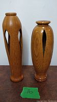 Front view of both tall and shorter wooden candle holders side by side on a wooden surface against a white wall.