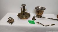 Five antique brass items arranged on white surface showing drawer pull, candle snuffer, footed dish, door knocker, and candle holder.