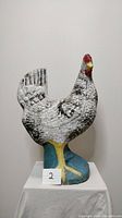 Front view of antique concrete rooster statue on a white cloth pedestal. The rooster is painted white with black feather details, red comb and wattles, yellow legs, standing on blue base.