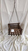 Wooden hanging planter suspended against a white draped background, showing front and side angle. Wooden slats form a square planter with metal chains attached for hanging.