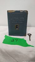Front view of antique Mutual Life of Canada dime-a-day coin bank showing textured green cover, embossed text, coin slot with coin counter, and original key.