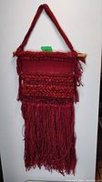 Full view of red hanging textile wall art showing overall shape, fringe, and wooden rod with strap