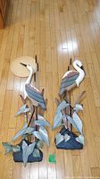 Front view of two large metal and wood hanging heron sculptures with foliage and rock bases on hardwood floor, one has a sun background piece.