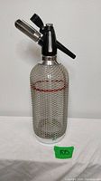 Full front view of the MCM seltzer/soda maker showing metal mesh wrap over clear glass bottle and black nozzle with silver metal lever.