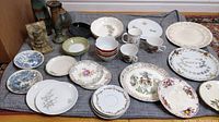 Overview of mixed collectible and antique plates, cups, decorative vases, stone and alabaster items on a grey mat.