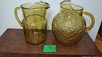 Pair of amber glass pitchers on brown surface with green lot number 90 visible, displaying both pitchers side-by-side.