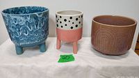 Photo showing all three ceramic planters on a white cloth background, including bright blue textured planter, pink with black polka dot planter, and dark pinkish planter with rainbow design.