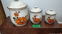 Set of three vintage ceramic canisters with orange/red poppy floral designs on a cream speckled base shown together on a wooden surface.