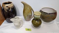 Five vintage ceramics including a dark brown jug with drip glaze, greenish-yellow pitcher, cream bird pitcher, green vase with lid, and planter bowl