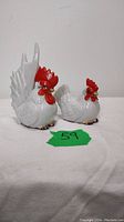 Porcelain rooster and hen figurines together on a white surface showing size relationship and overall condition.