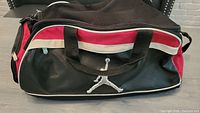 Front view of the large black gym bag with red and white detailing and partially lifted white Jumpman logo on the pocket.