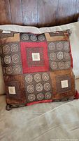 Colorful decorative pillow resting on a couch, showing a detailed patchwork design with squares and swirls in earth tones and red accents.