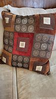Colorful decorative pillow positioned on beige couch showing geometric patchwork design with spirals and squares in warm earth tones.
