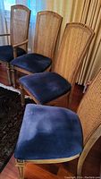 Four light brown wood dining chairs with cane backs and blue velvet cushions. Two of the chairs have armrests, two do not. Visible wood grain and slightly distressed finish.