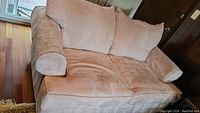 Three quarter angled view of a light golden beige microfiber love seat placed on a hardwood floor near French doors showing armrest, cushions, and seat upholstery.