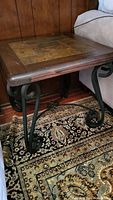 Side view of the rustic side table showing wood frame, stone tile top, and scroll-style black iron legs resting on a patterned rug.