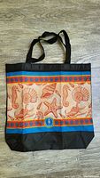 Front view of beach tote bag showing sea-themed vinyl print with seahorses, shells and starfish in orange on a light background bordered by blue and black.
