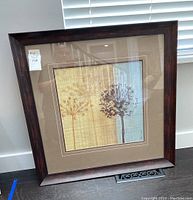 Frontal view of framed art print resting on floor against wall, showing full wood frame and botanical artwork.