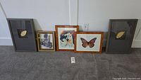 Five framed art pieces on floor including two shadow boxes with gold leaf, a horse portrait, a monarch butterfly print, and one abstract artwork