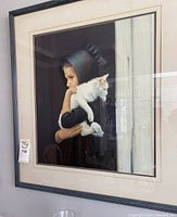 Framed art showing a woman in a dark hood holding a white cat, with reflections visible on the glass surface.