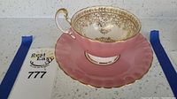 Side view of the Aynsley pink and gold teacup sitting on its matching saucer with scalloped edges and gold trim.